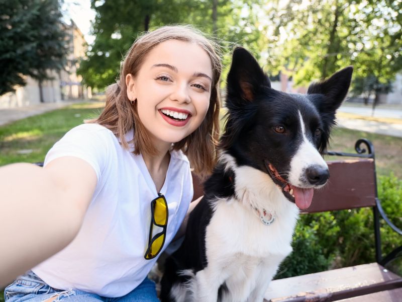 Young woman in white shirt taking a selfie with a happy black and white dog on a bench in a sunny park.