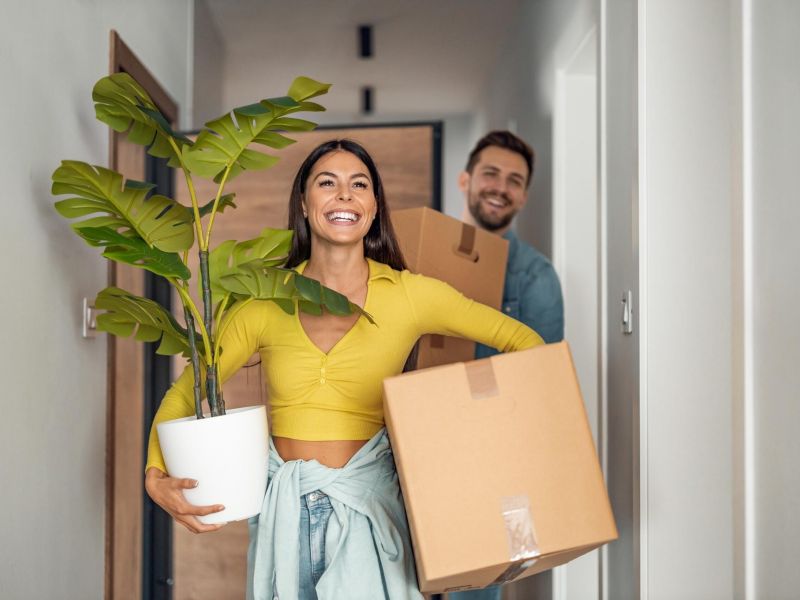A smiling couple walking into their new home carrying cardboard boxes and a potted plant.