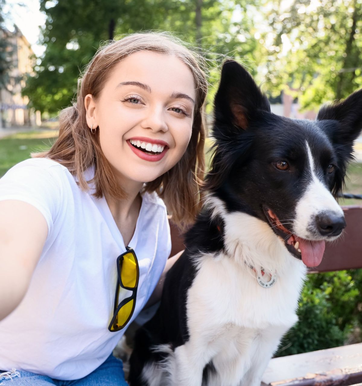 Young woman in white shirt taking a selfie with a happy black and white dog on a bench in a sunny park.